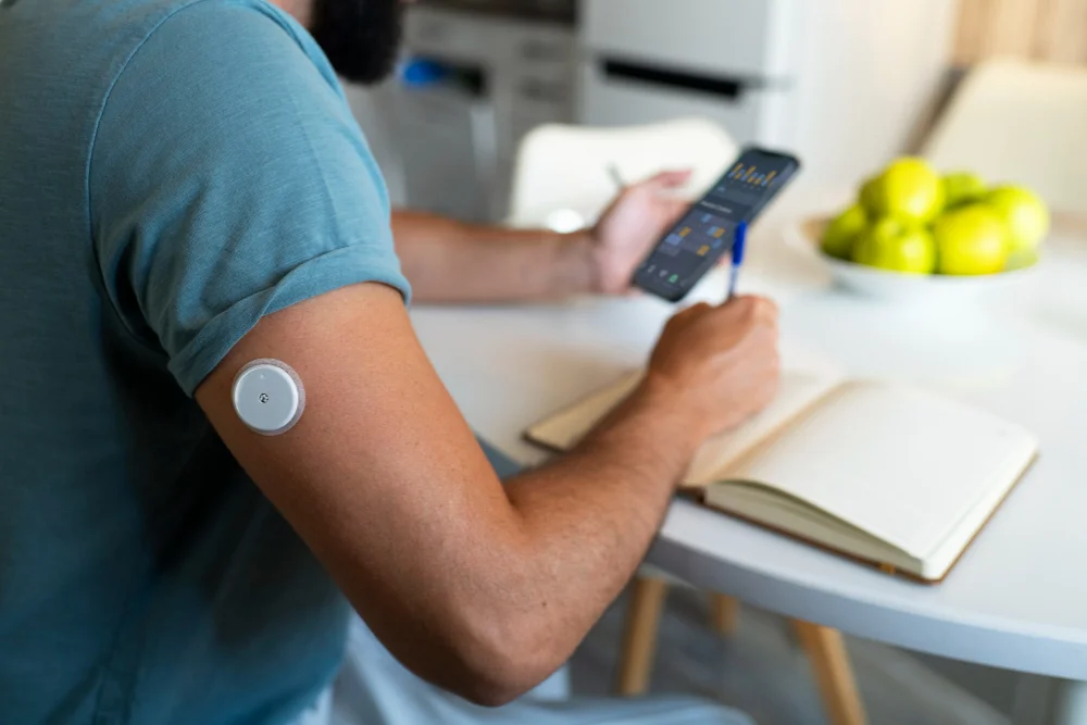 Man with a continuous glucose monitor in his right upper arm checking statistics on his phone while writing in a book.