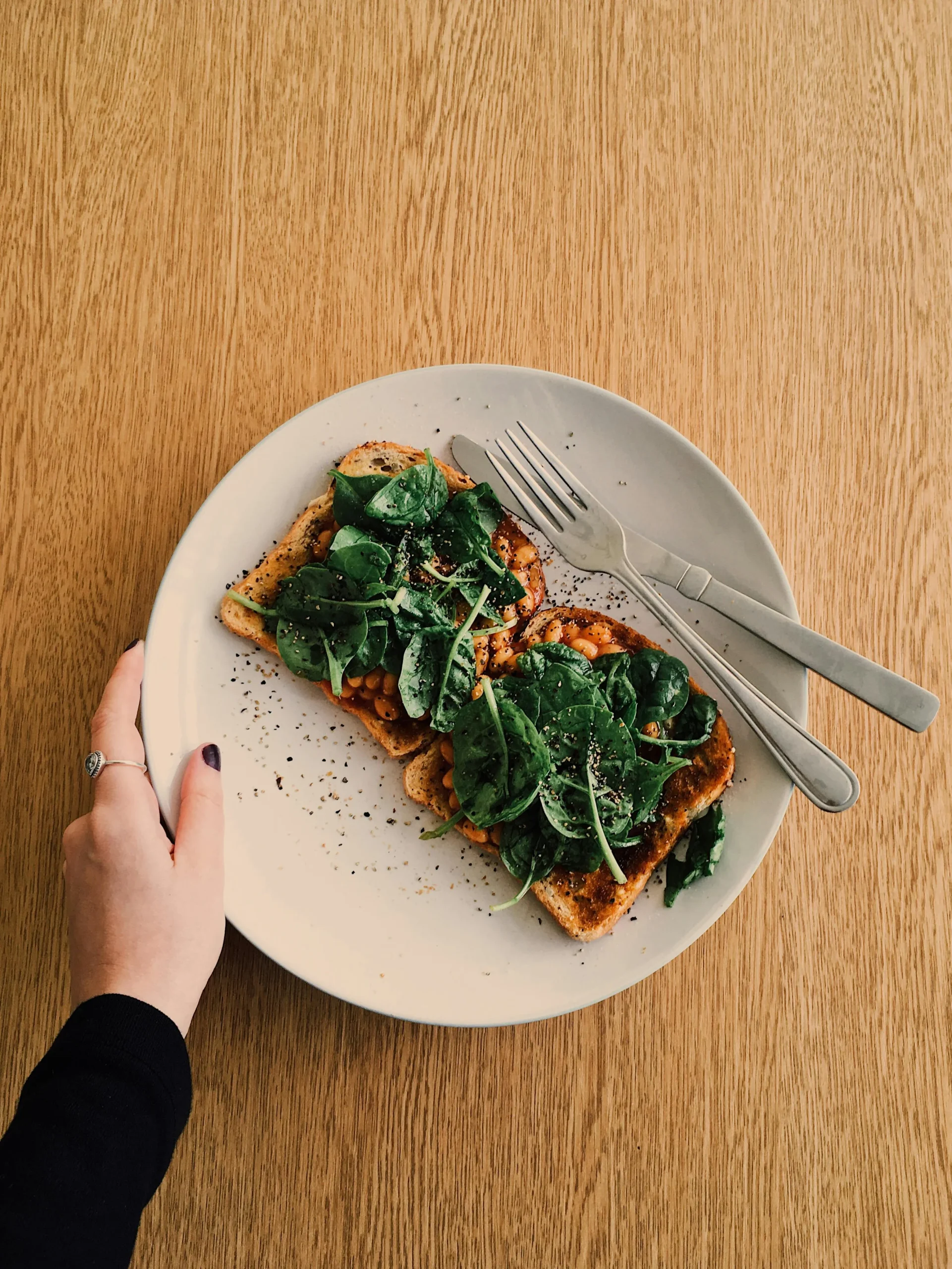 A hand holding a white plate with cutlery, bread with beans and vegetables