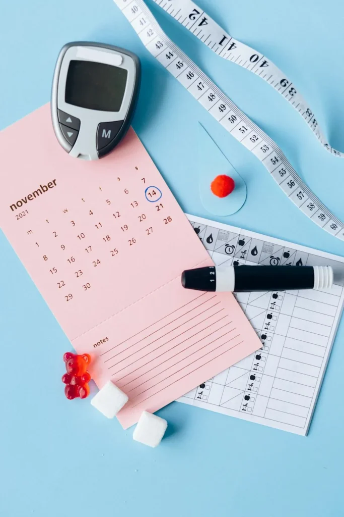 Resting on a light blue background is a glucometer, white tape measure, blood lancet pen, a pink calendar of November 2021, sugar cubes, and a red gummy bear