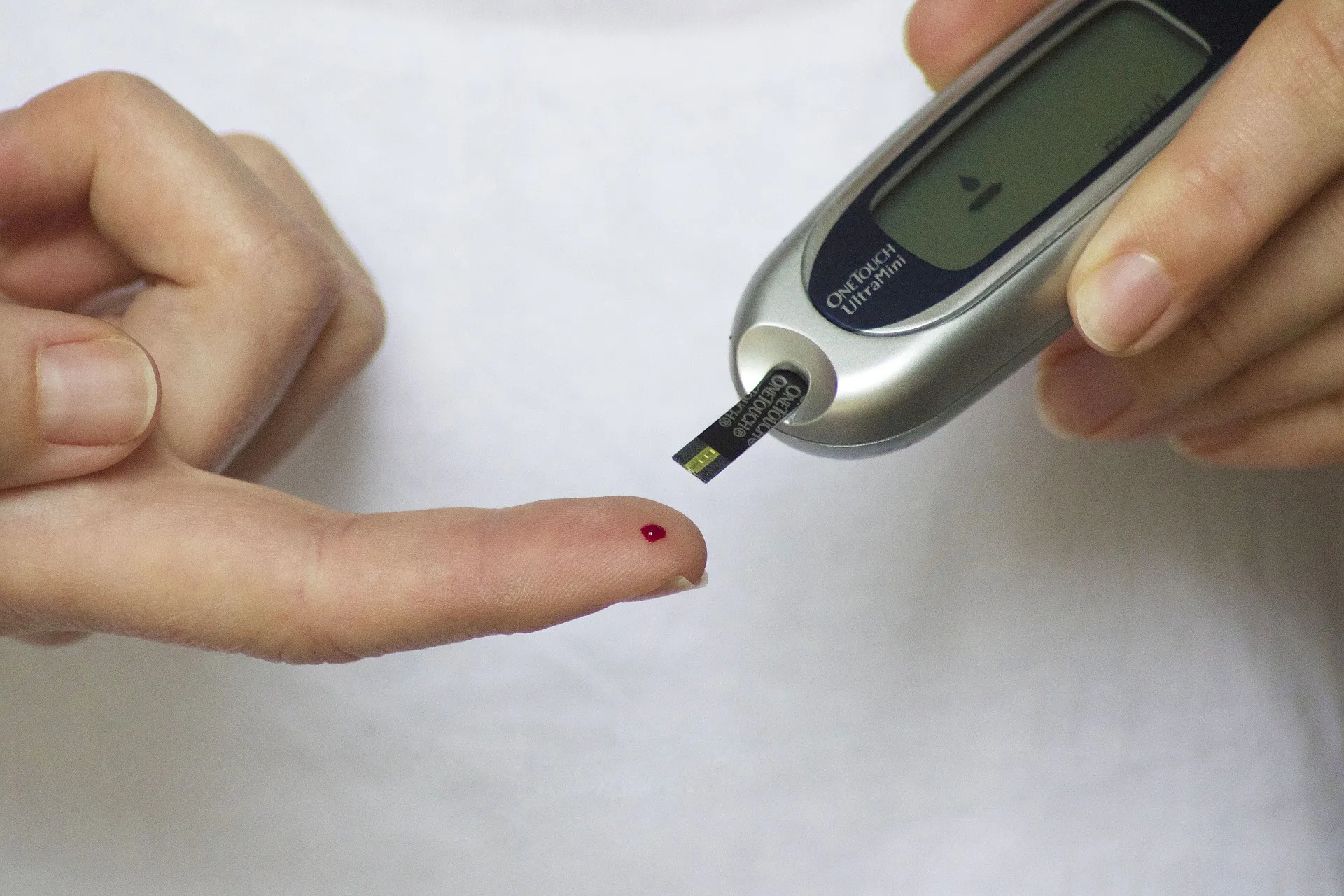 A person with a blood drop on the index finger of one hand and a glucose meter loaded with a test strip in the other hand.