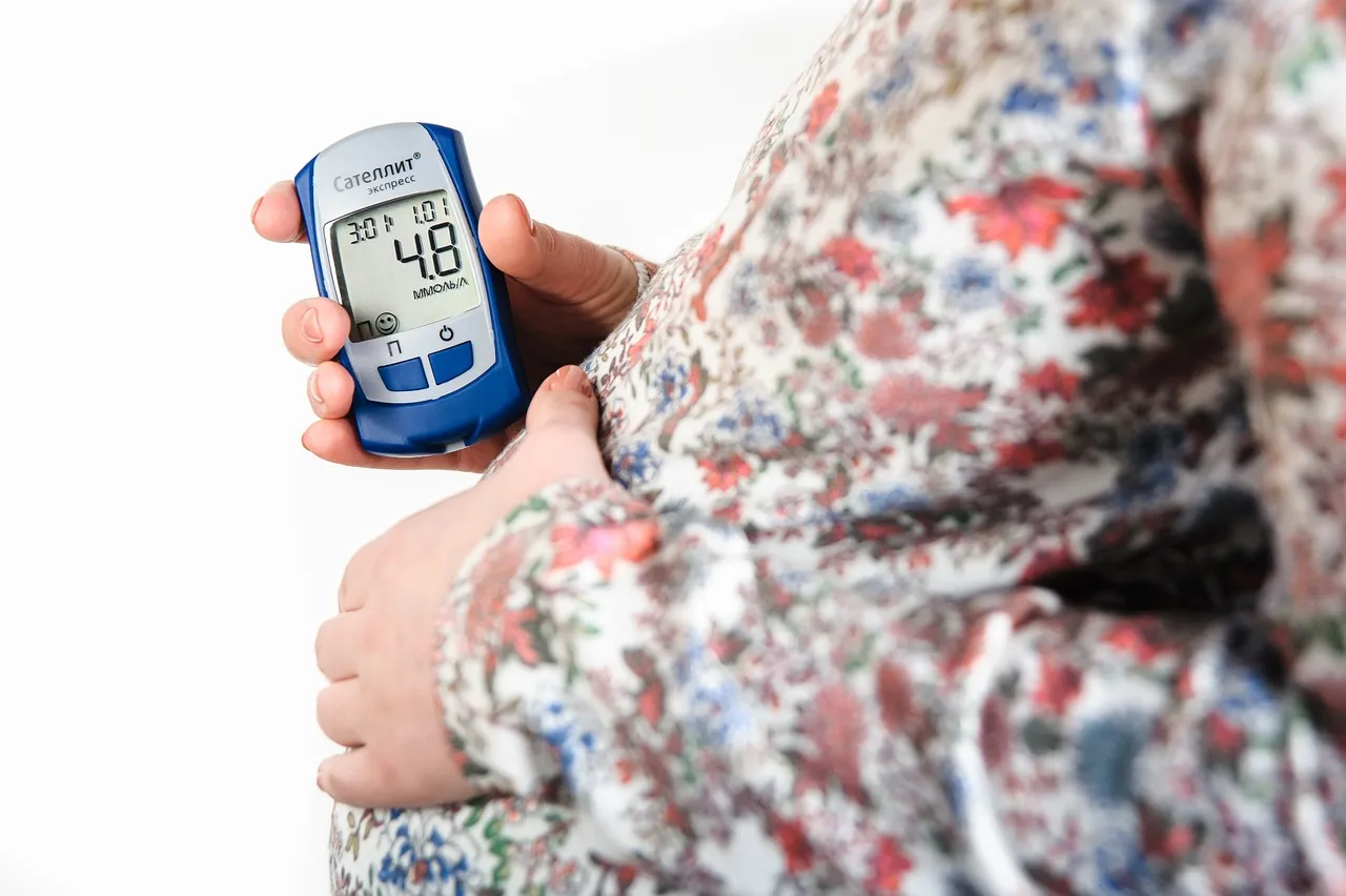 A pregnant woman wearing a floral dress, touching her belly with her left hand and holding a glucometer in her right hand.