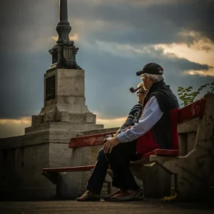 Elderly male with a smoking pipe in his mouth seated on a bench beside an elderly woman.