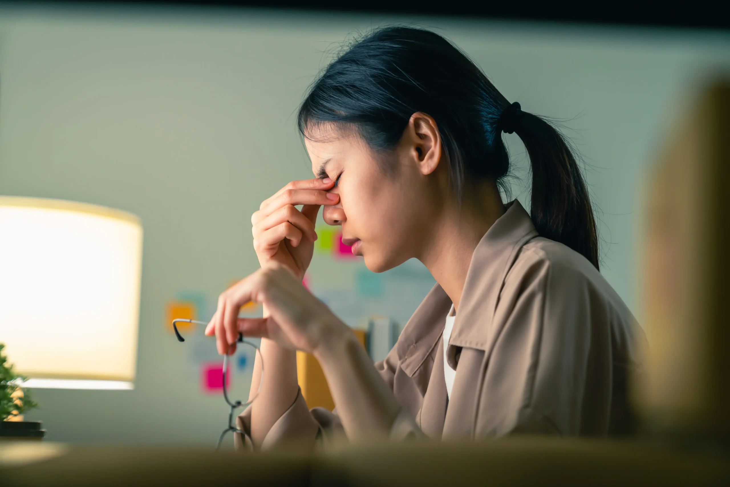 An Asian woman holding eyeglasses in her left hand and touching her closed eyes with the right hand